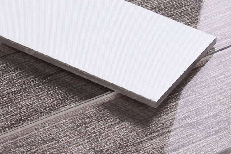 Close-up of a white tile sample on a textured grey wood-like floor, showcasing installation materials.